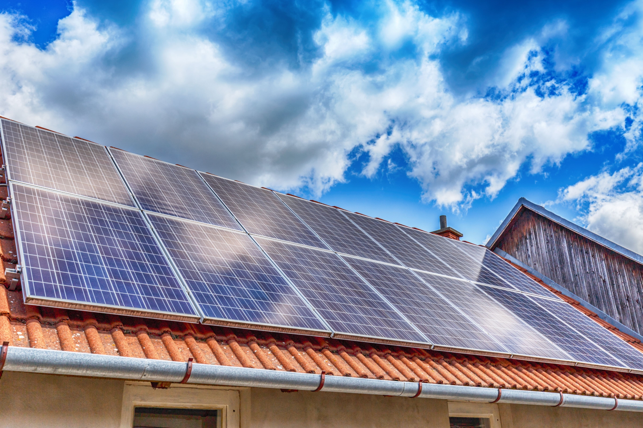 Large array of solar panels mounted on a red terracotta tiled roof under a bright, cloudy sky.