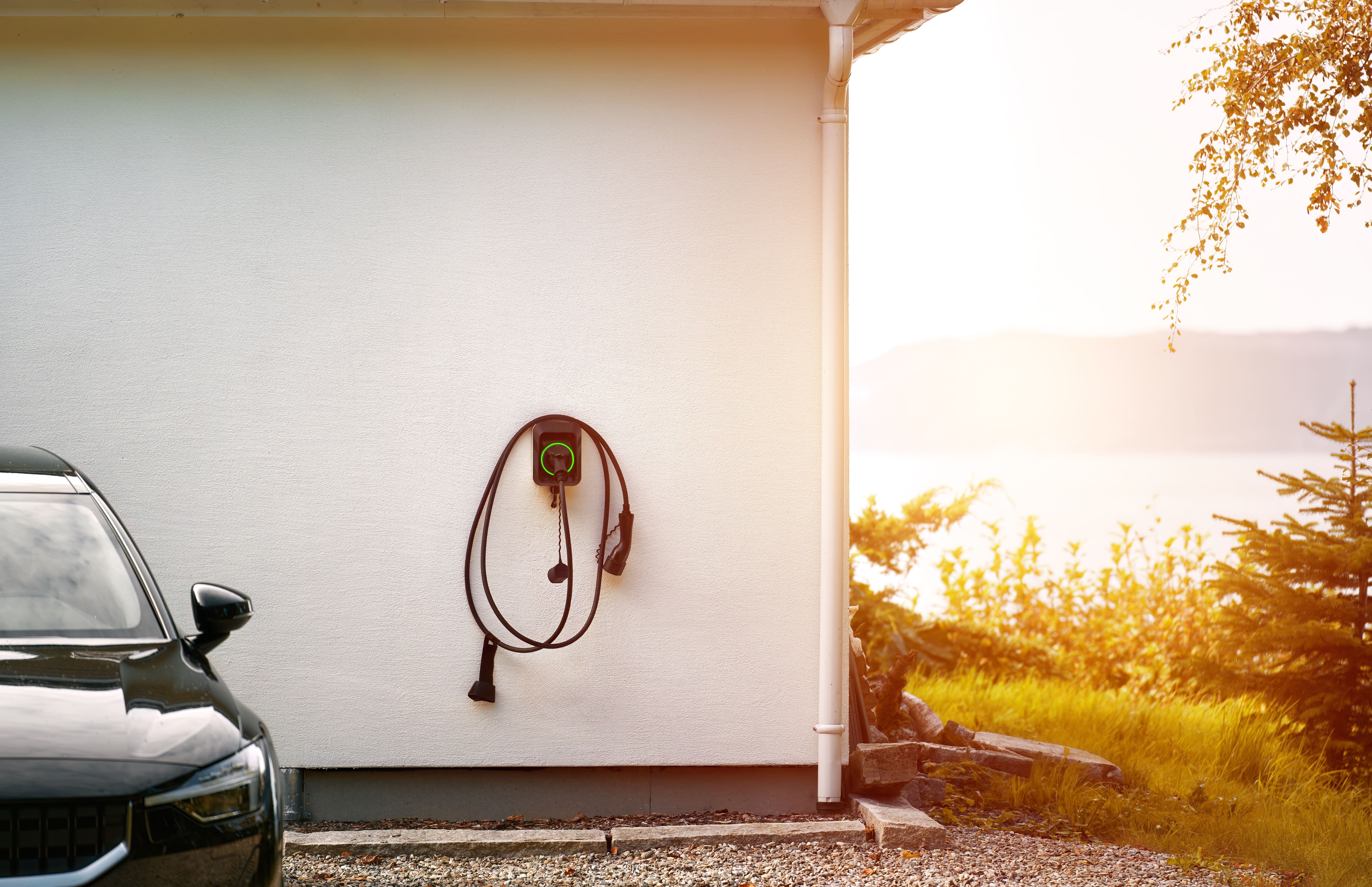 AdobeStock_663678264 (1) An electric vehicle charging point mounted on a white exterior wall next to a black car, with a scenic lake and sunset in the background.
