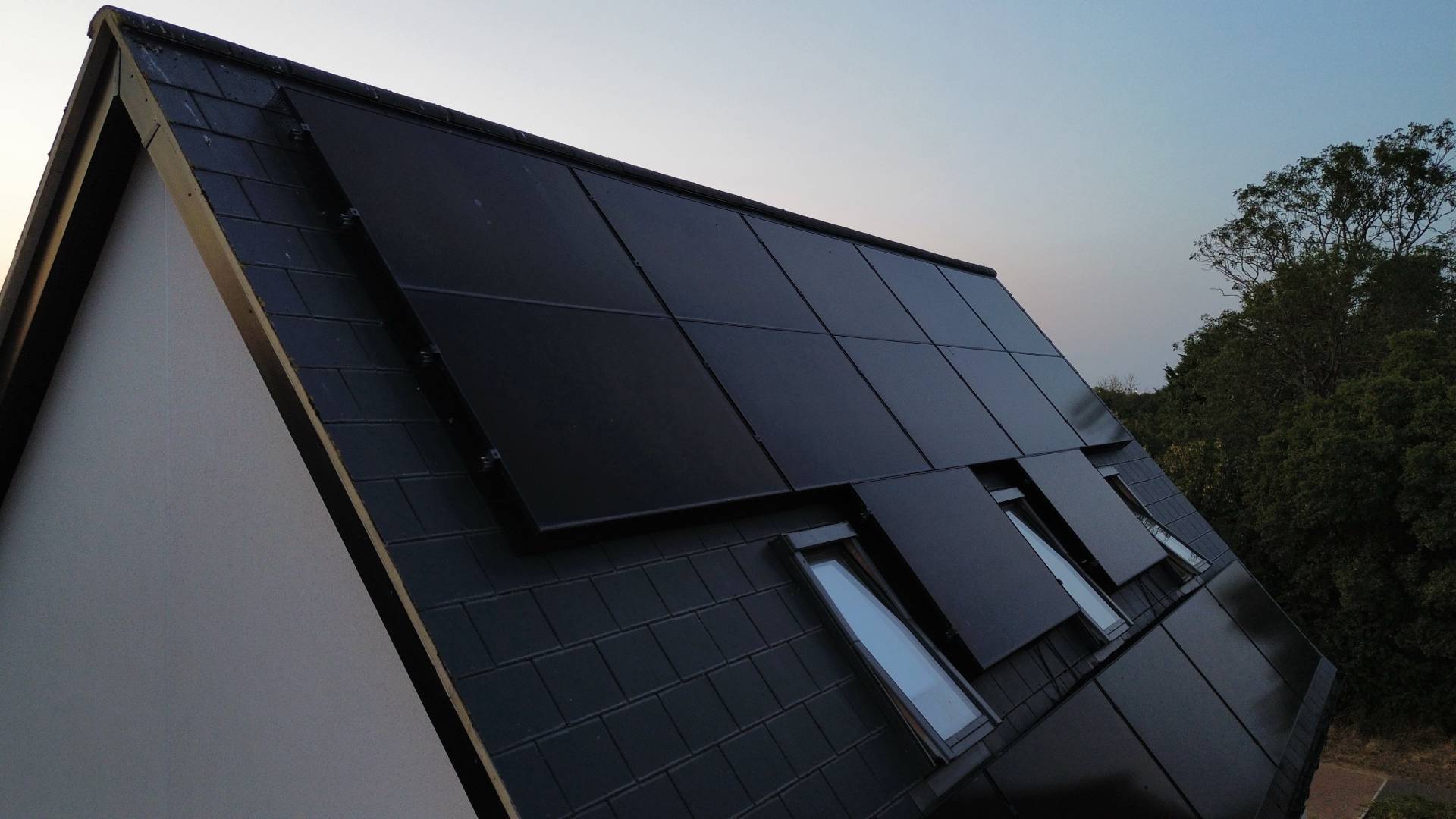 Angled view of slate roof with black integrated solar panels fitted above three roof windows on a modern home at dusk