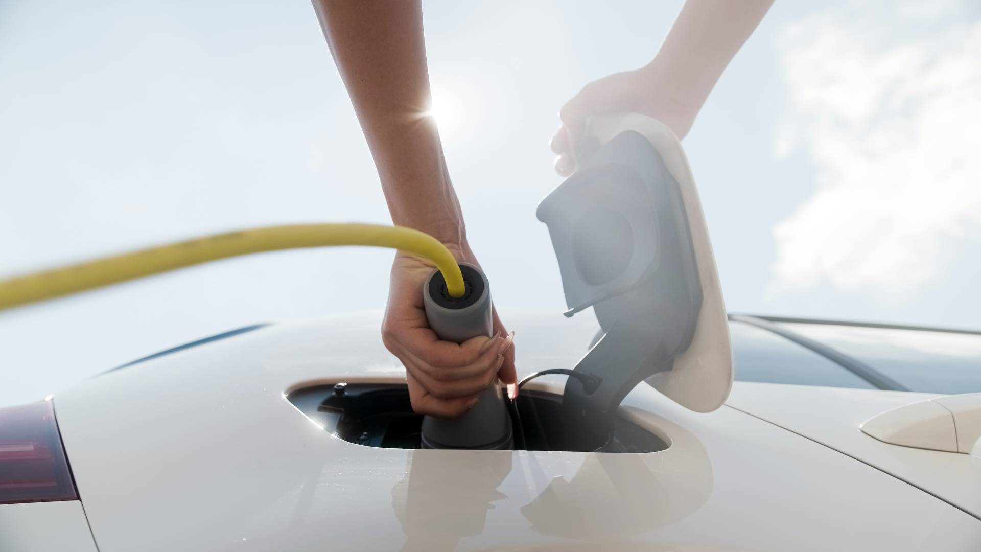AdobeStock_453178326 Person plugging electric vehicle charging cable into EV under bright sunlight