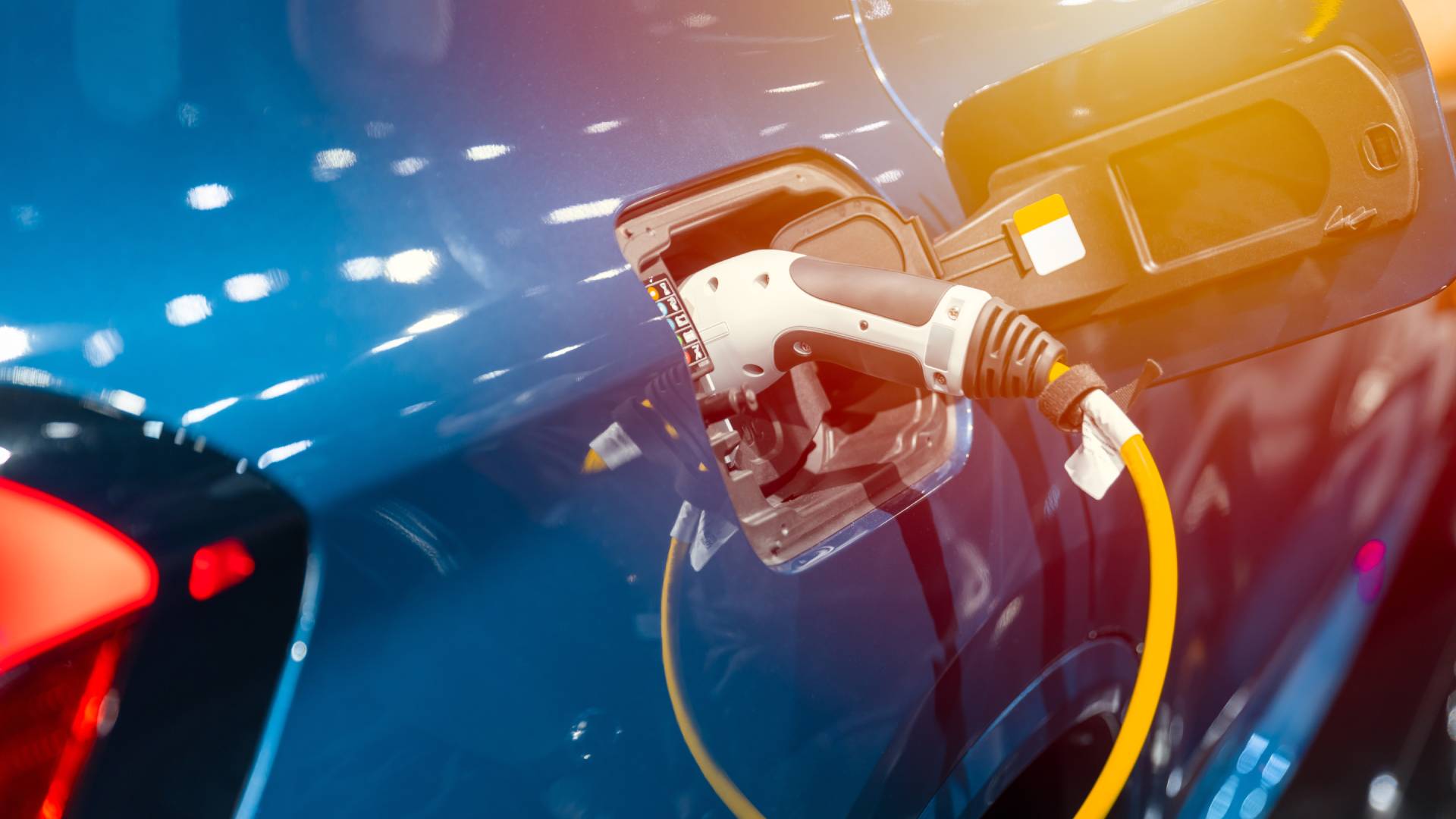 AdobeStock_496988767 Electric vehicle charging cable plugged into blue EV at charging station with yellow lead and lens flare lighting