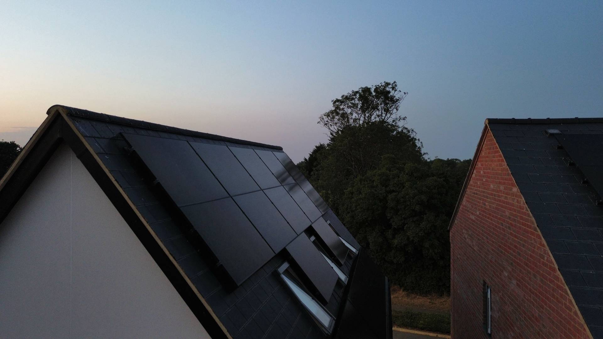 Black in-roof solar panel system on pitched slate roof with roof windows at dusk beside neighbouring brick house