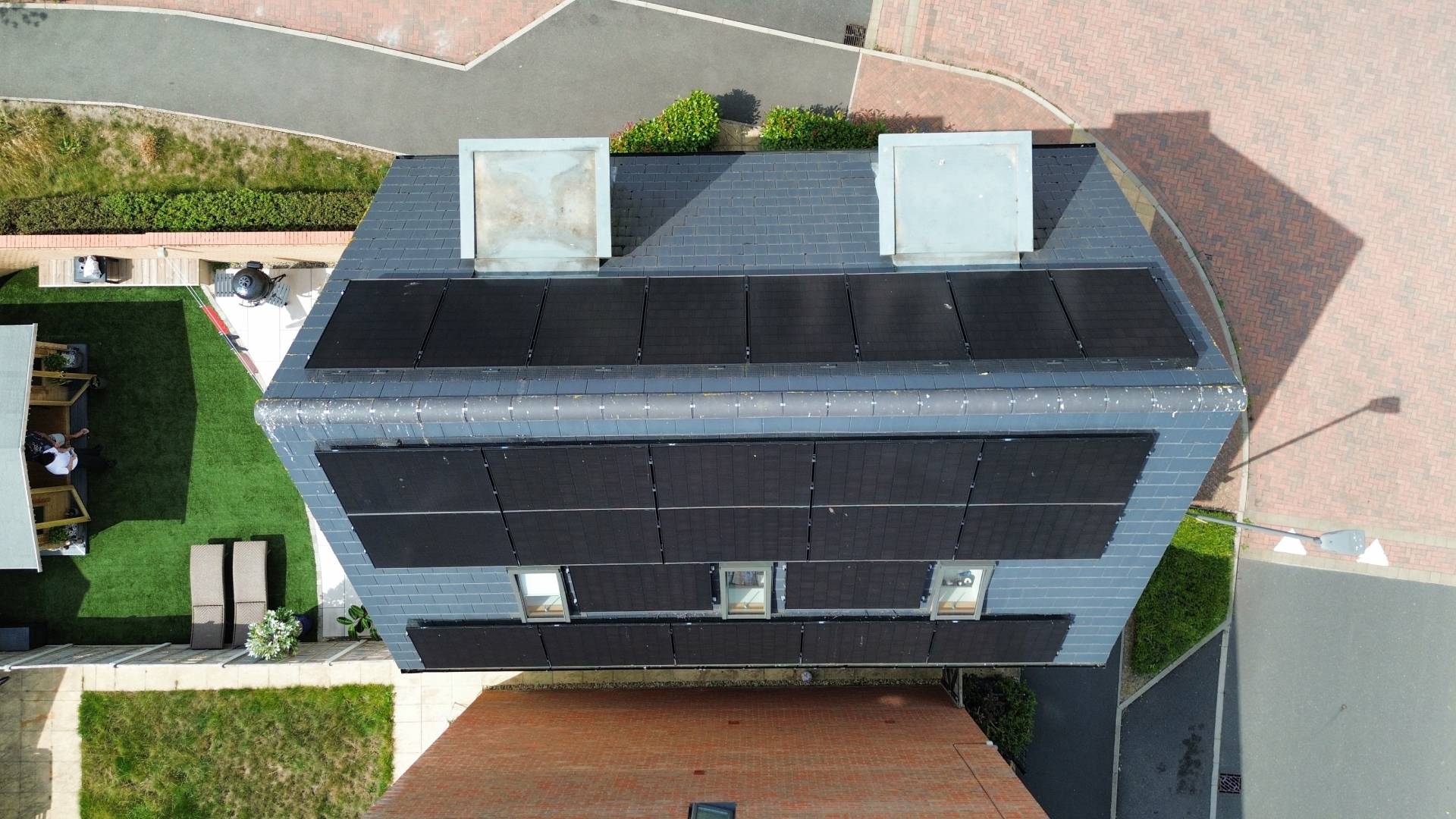 Top-down aerial view of a modern two-storey home with multiple black solar panel arrays installed across the main roof sections, set within a neatly landscaped garden and paved driveway in a residential neighbourhood.