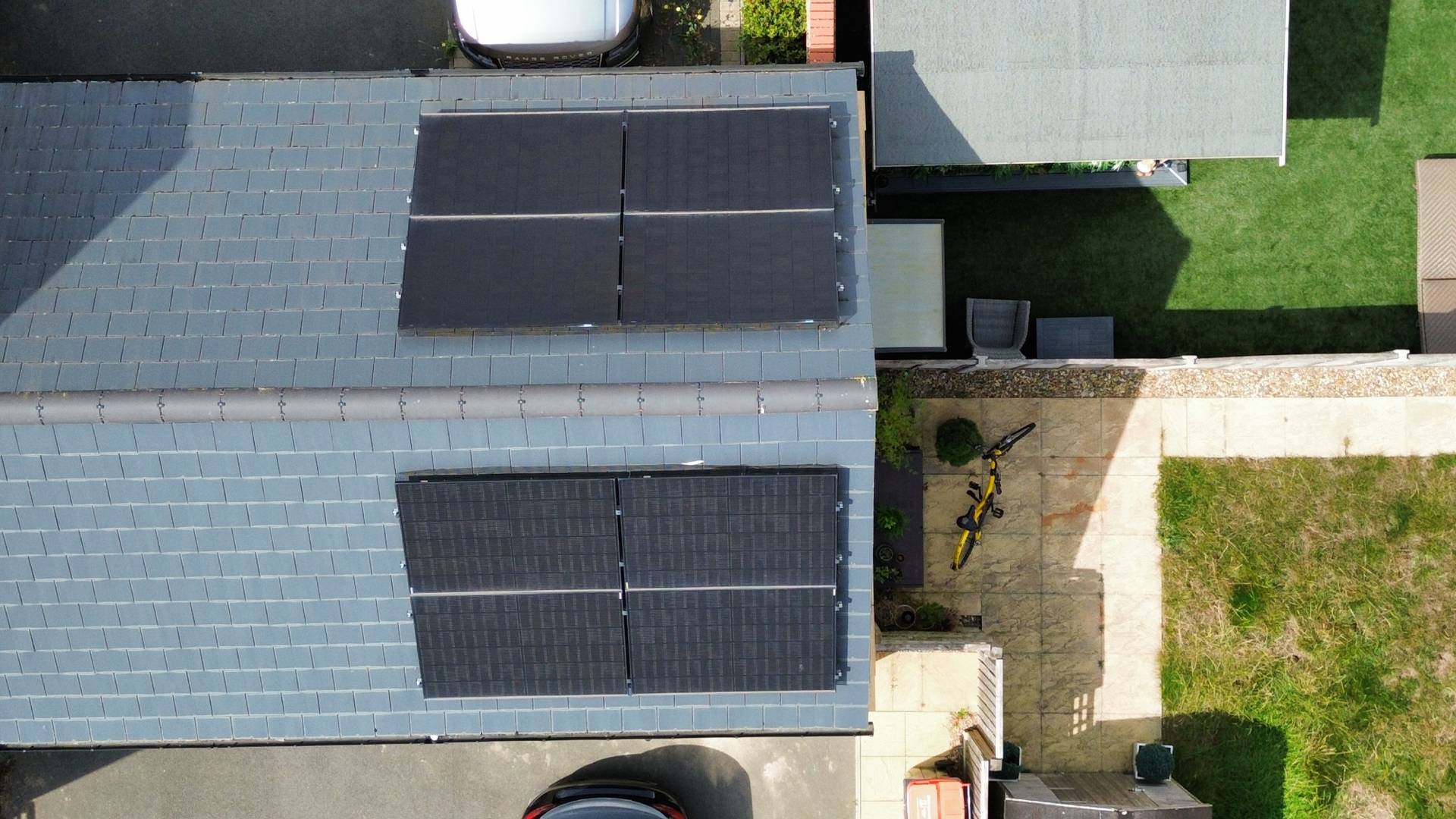 Top-down aerial view of a modern residential property with multiple solar panel arrays installed on the main house and garage roofs, featuring a landscaped garden, patio area with outdoor furniture, and parked cars along the street.
