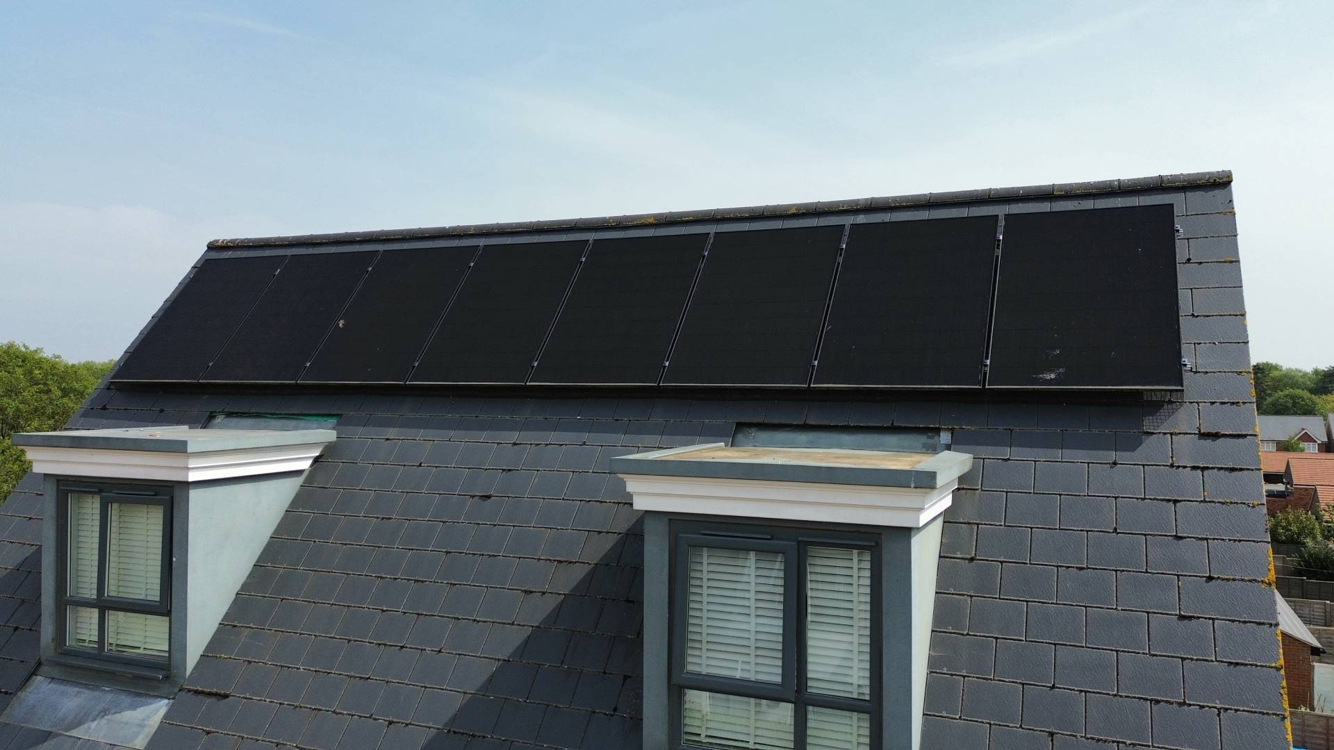 Close-up of black solar panels installed on a slate-tiled residential roof, mounted in a neat row above two dormer windows on a modern home.