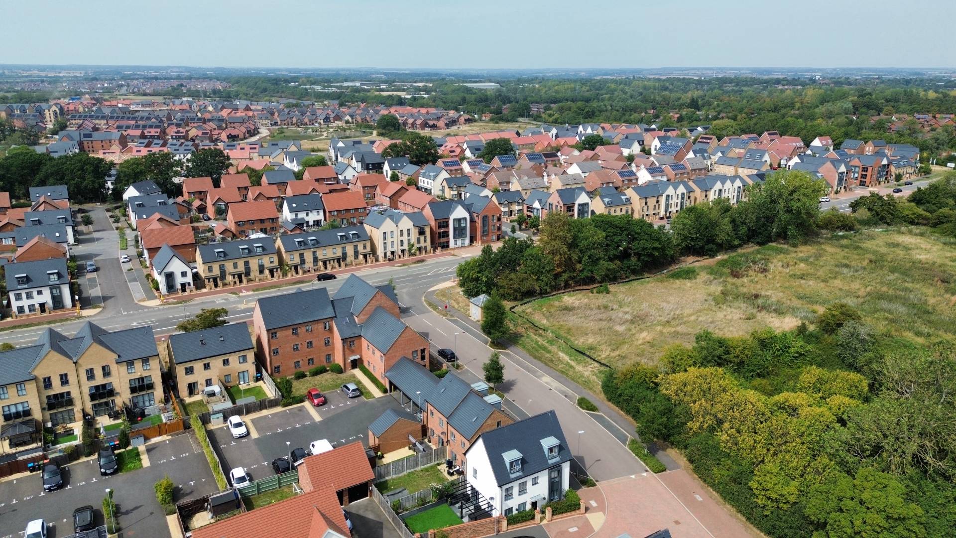 Aerial daytime view of a modern residential development with rows of new-build houses, red and grey roofs, tree-lined streets, and open green space bordering the estate.