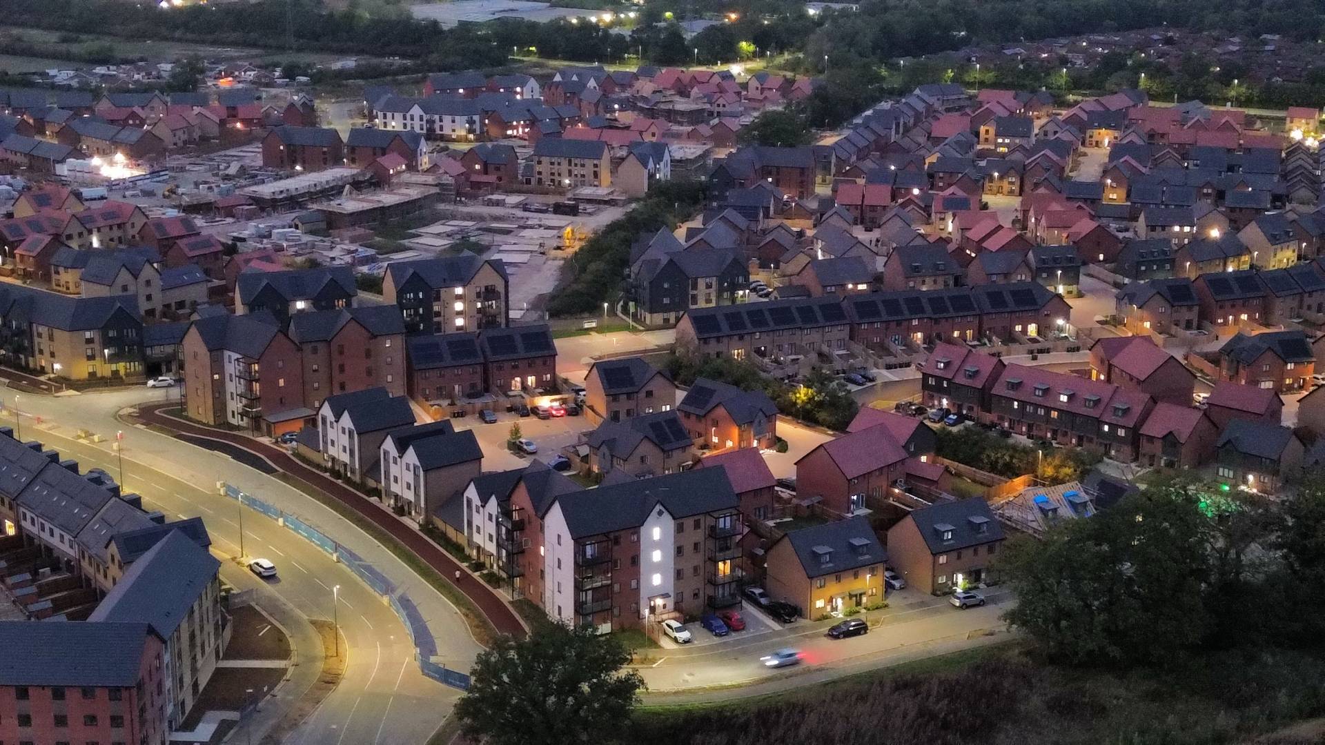 dji_fly_20250813_203248_55_1755165917518_photo_optimized (1) Aerial evening view of a modern housing development with rows of newly built homes, apartment blocks, and illuminated streets, with ongoing construction visible in the background.