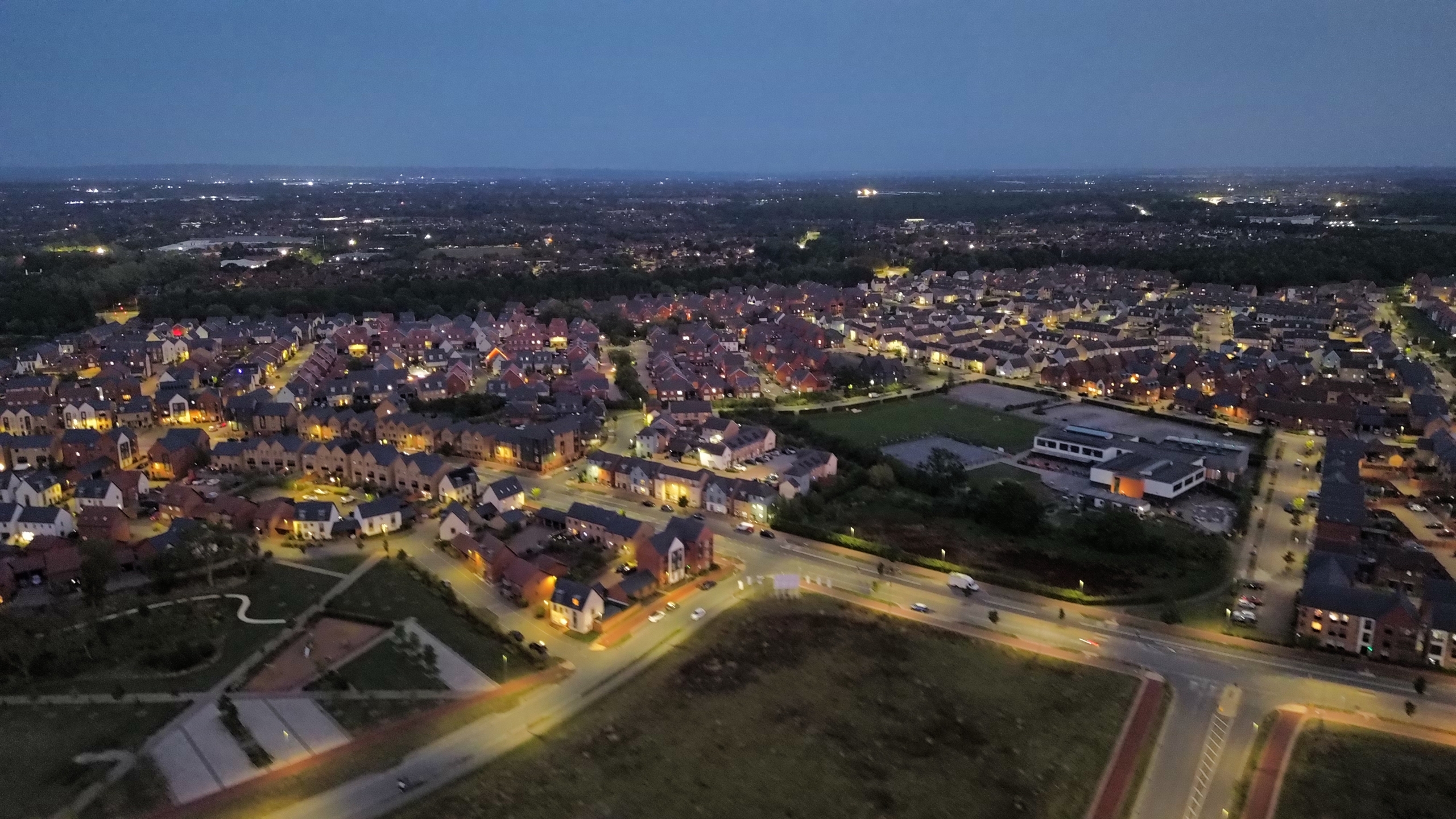 DCIM100MEDIADJI_0061.JPG Aerial dusk view of a residential neighbourhood with rows of houses, lit streets, and surrounding greenery stretching into the distance.