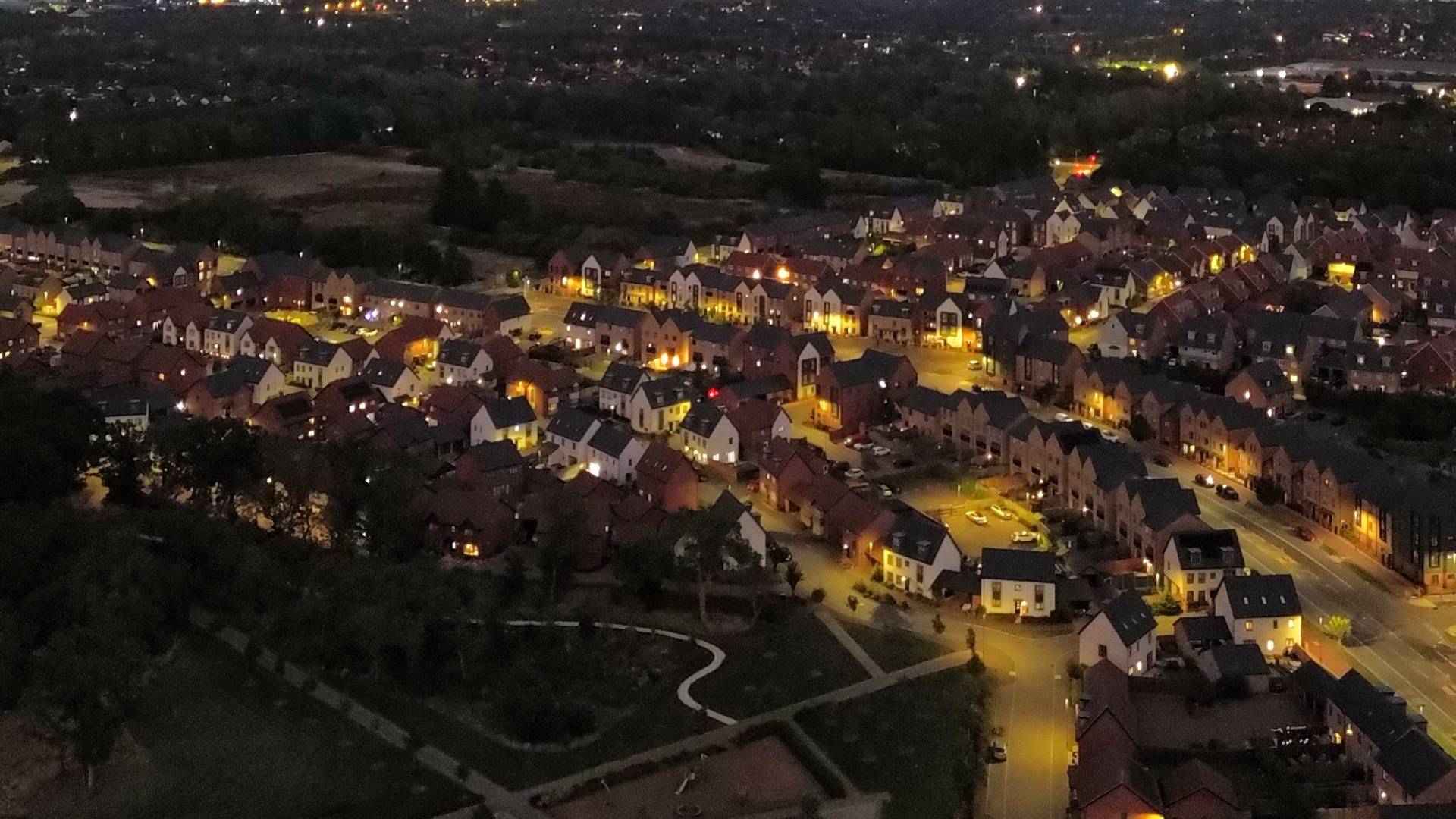 Aerial view of a residential neighbourhood at night, with houses and streets lit by warm streetlights and surrounding countryside in darkness.