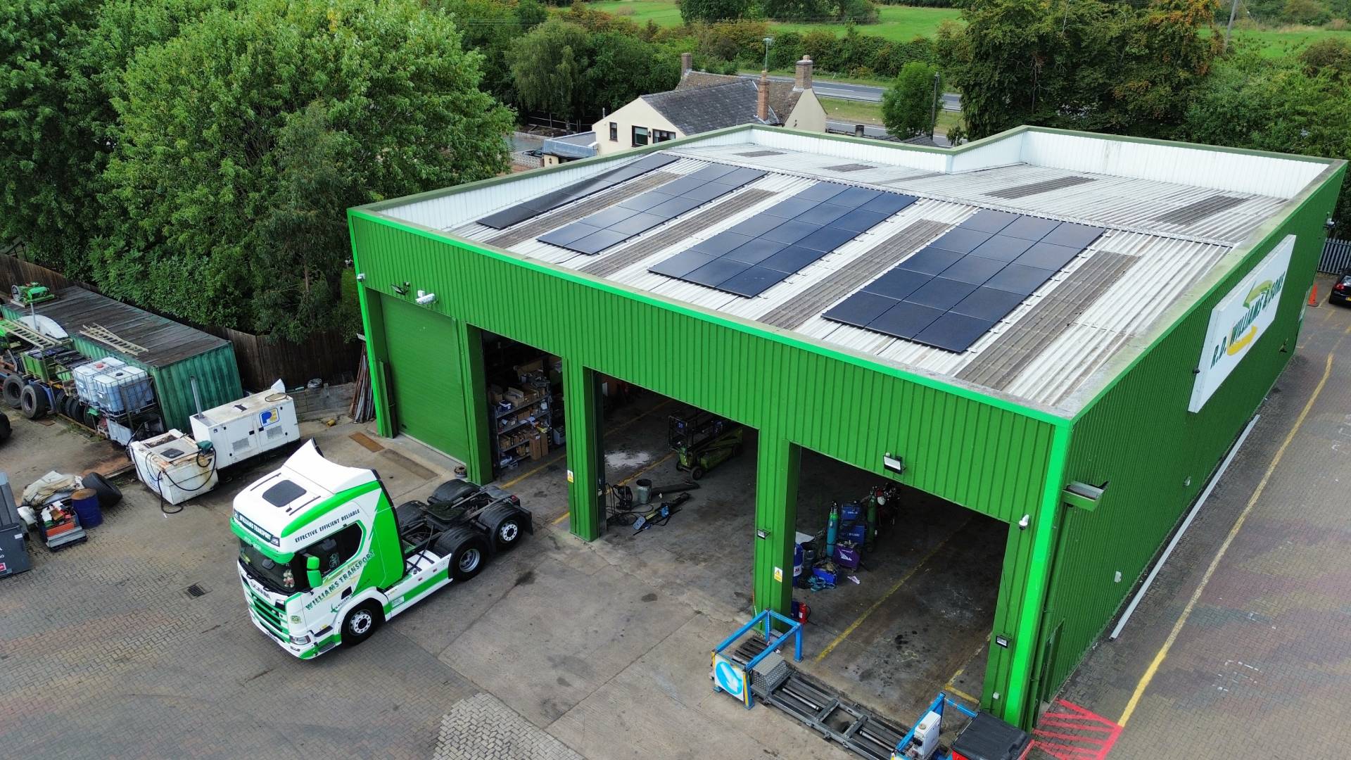 Drone view of a green industrial warehouse fitted with multiple rows of rooftop solar panels, with large open workshop bays below and a white and green lorry cab parked in the yard beside the building, surrounded by trees and nearby houses.