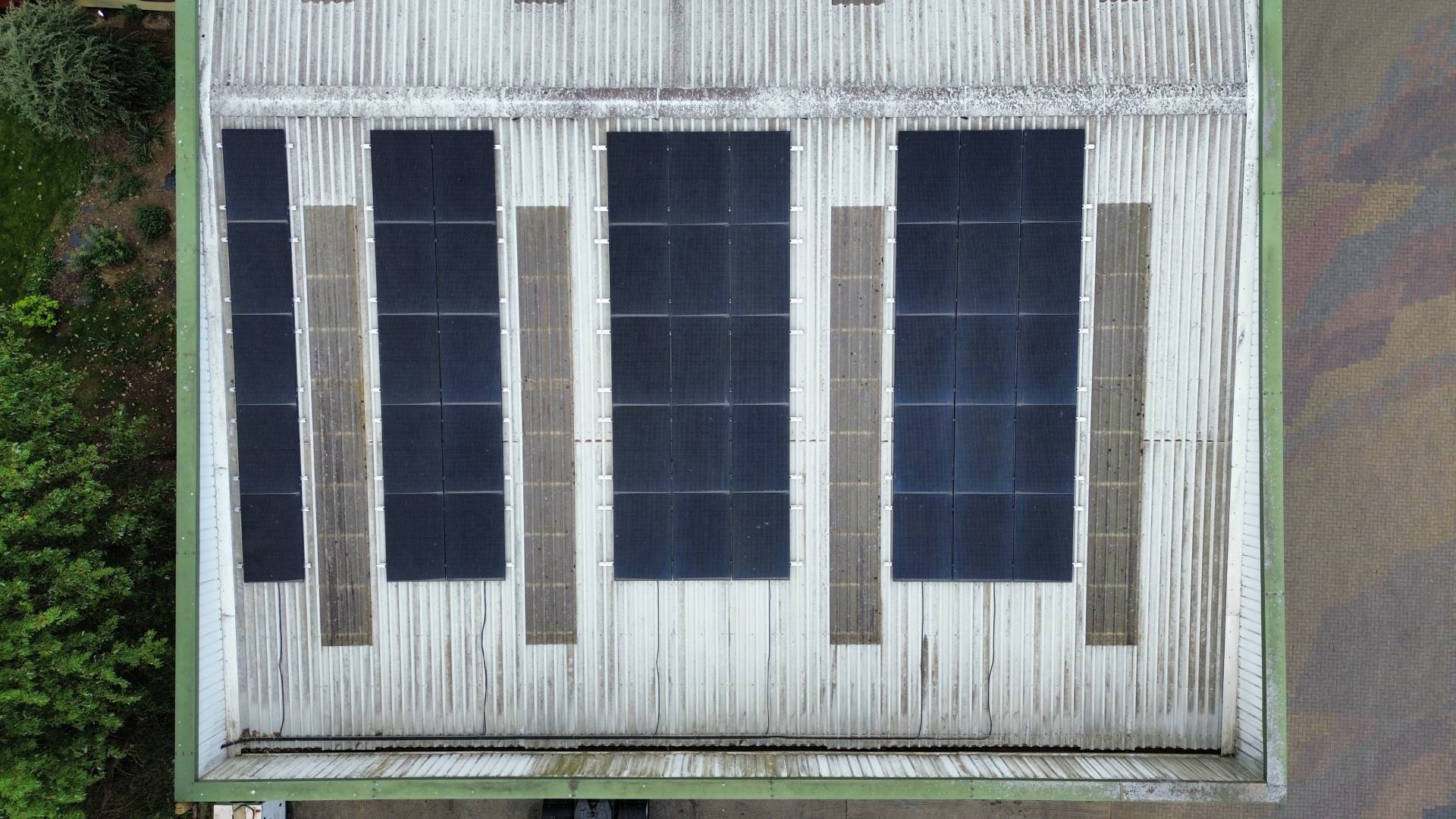 Aerial top-down view of a corrugated metal roof fitted with multiple rows of black solar panel arrays, evenly spaced across the surface.