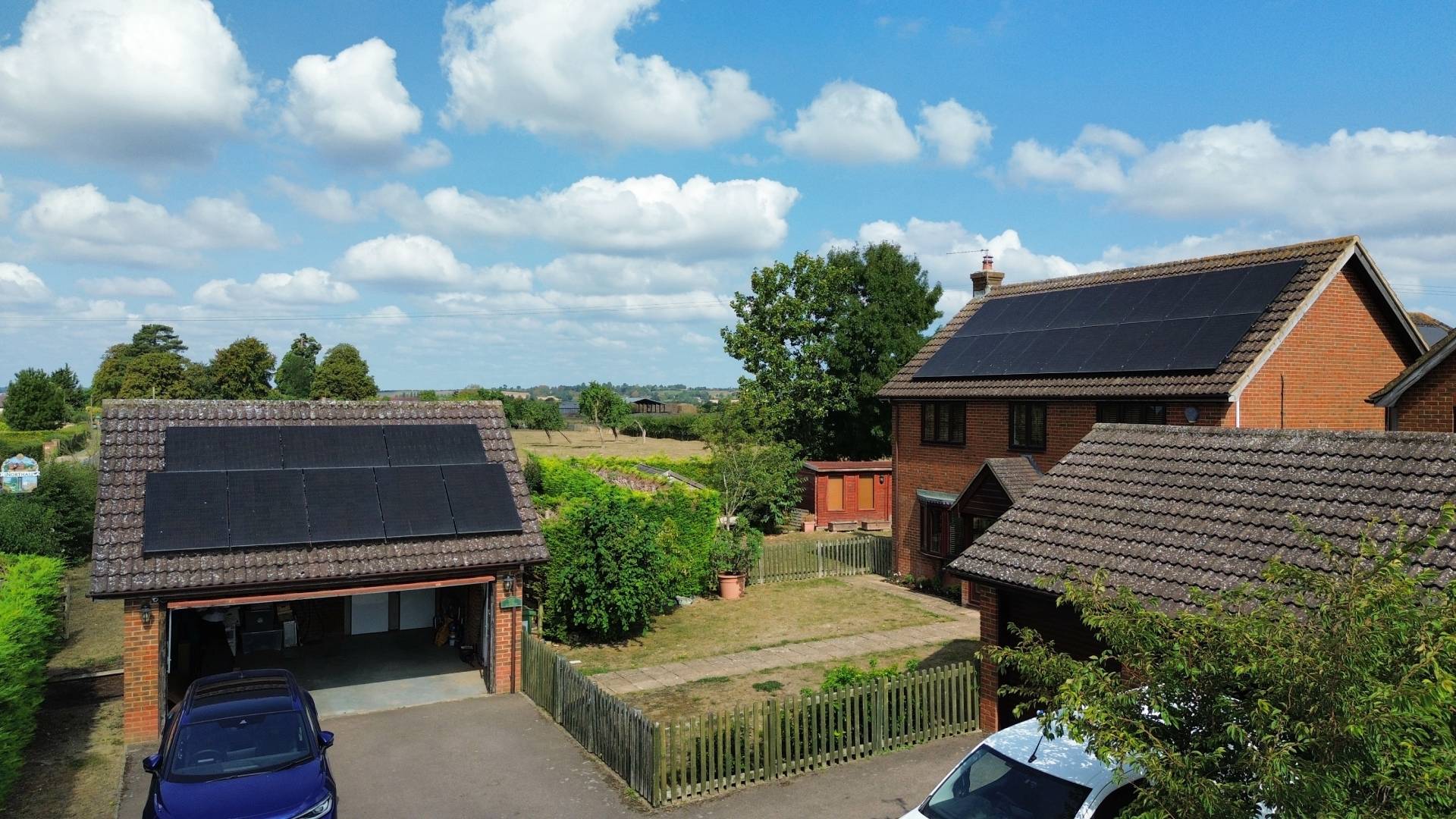 dji_fly_20250815_103826_184_1755263089627_photo_optimized (1) Detached brick home and separate garage fitted with black solar panel arrays on both roofs, viewed from an elevated angle on a bright day. The property features a driveway with parked cars, a fenced garden, and open countryside in the background under a blue sky with scattered clouds.